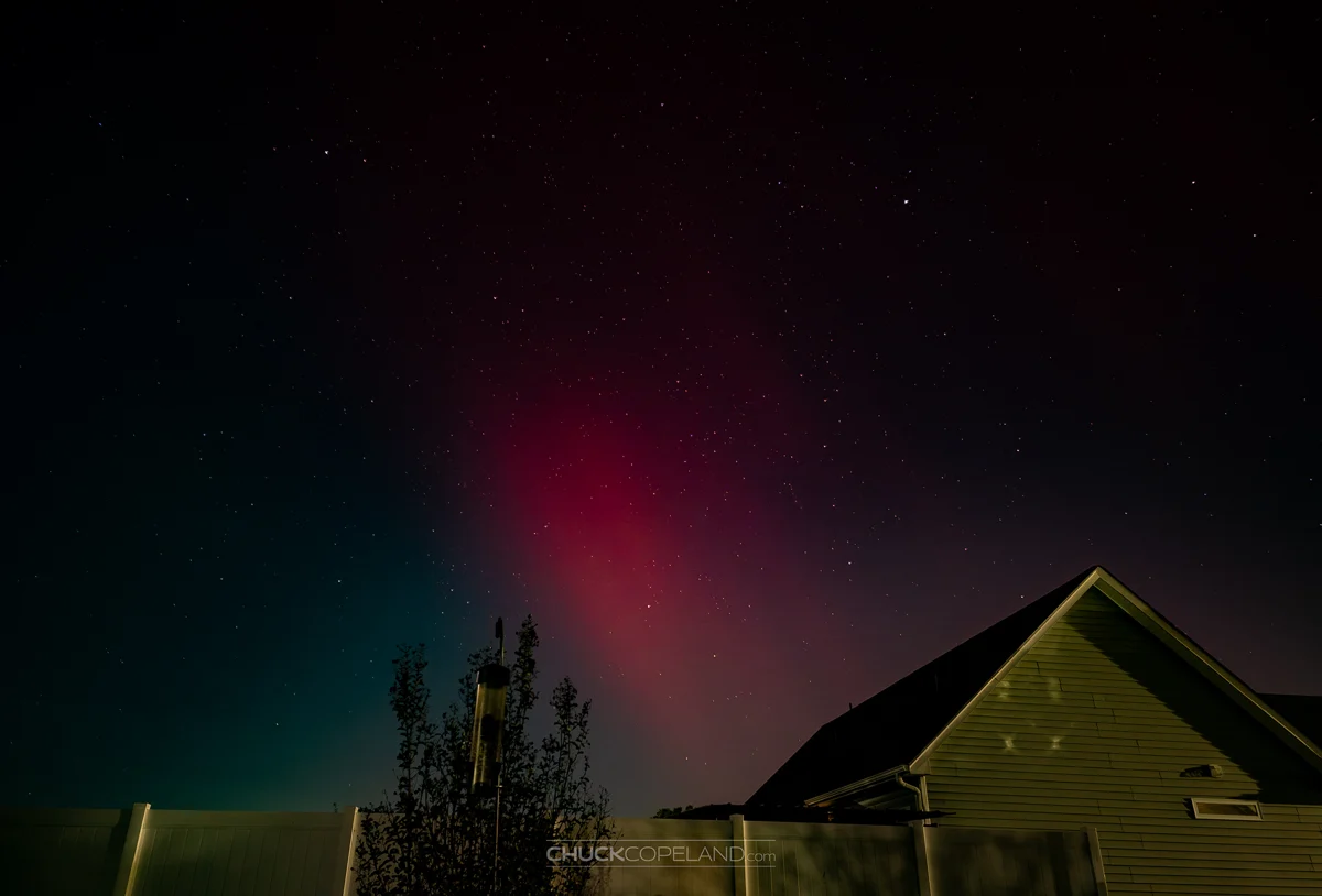 Pink and green aurora glowing above a house and star-filled sky